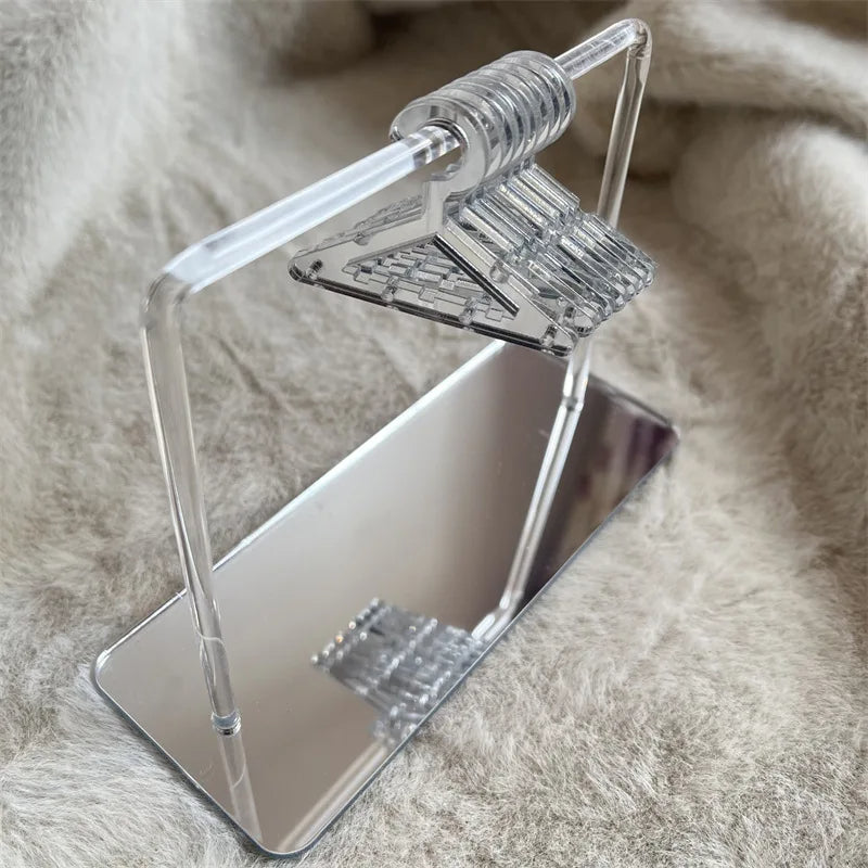 Rangement pour bijoux argenté avec cintres transparents sur socle miroir, présenté dans une ambiance minimaliste et raffinée.